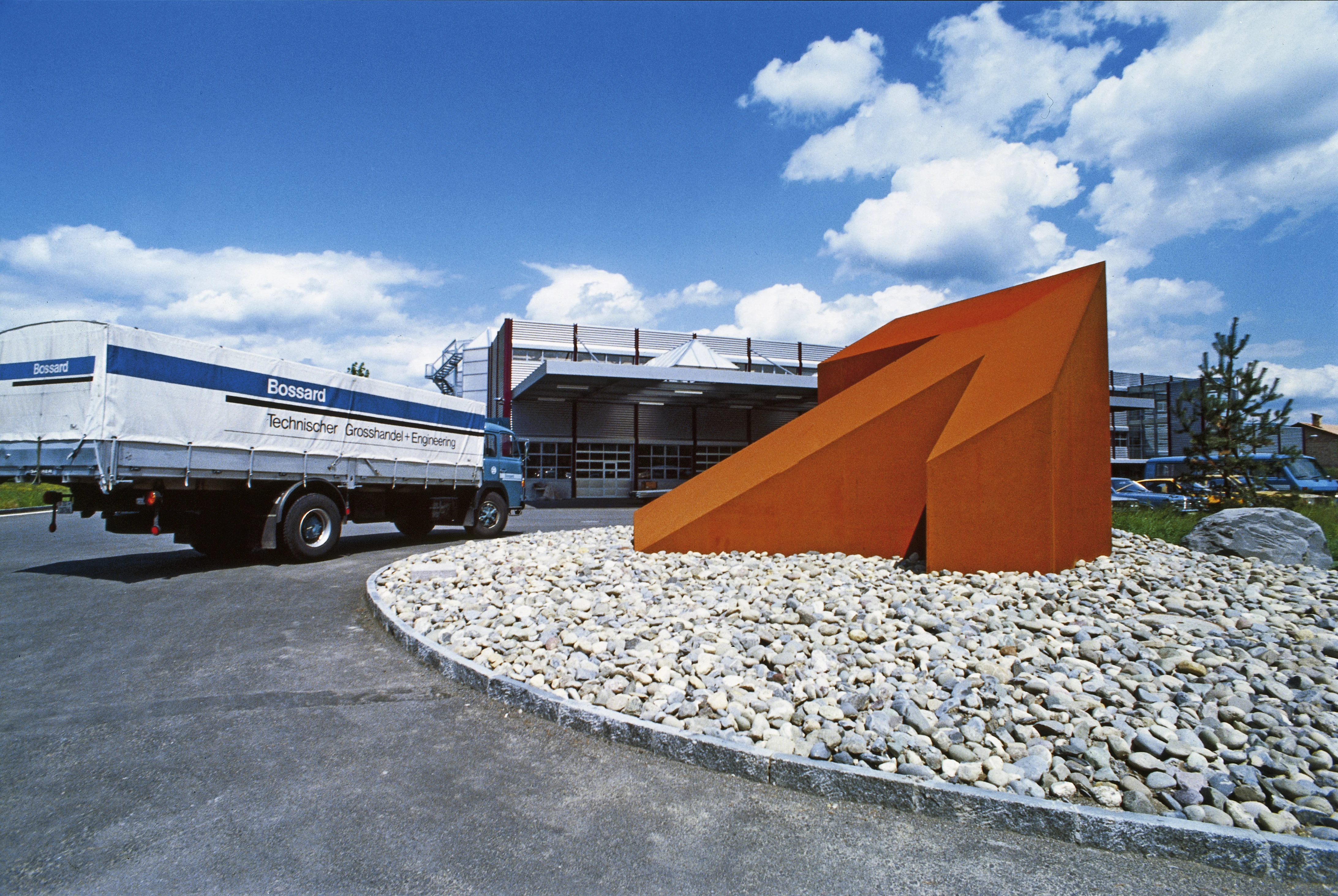 Historisk billede af en stor orange pilskulptur i en rundkørsel, med en Bossard-lastbil og lager i baggrunden.