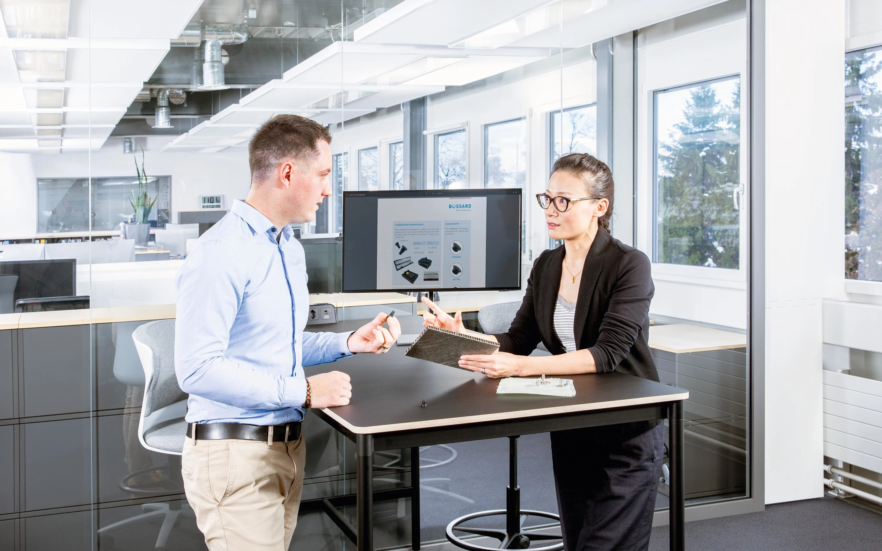Two people at a desk in a modern office discuss fastening a composite material; one holds a sandwich panel, the other a fastener.