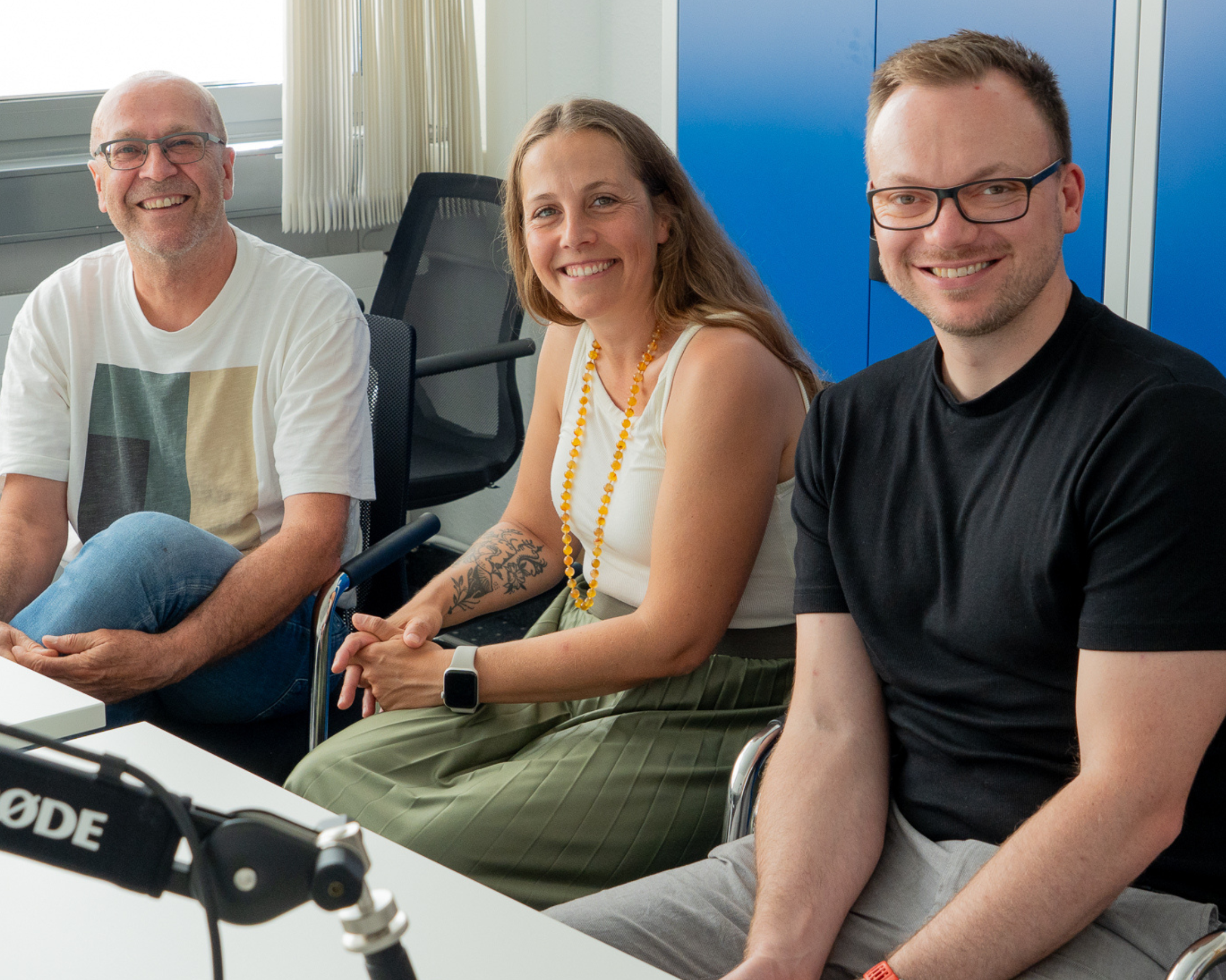 Walter Scharpf, Maja Matzke und Tobias Engelberg sitzen lächelnd an einem Tisch bei einer Podcast-Aufnahme; im Vordergrund steht ein Mikrofon.