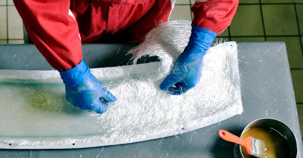 Person in roten Arbeitskleidung und blauen Handschuhen verarbeitet Glasfasermatten auf einer Form.