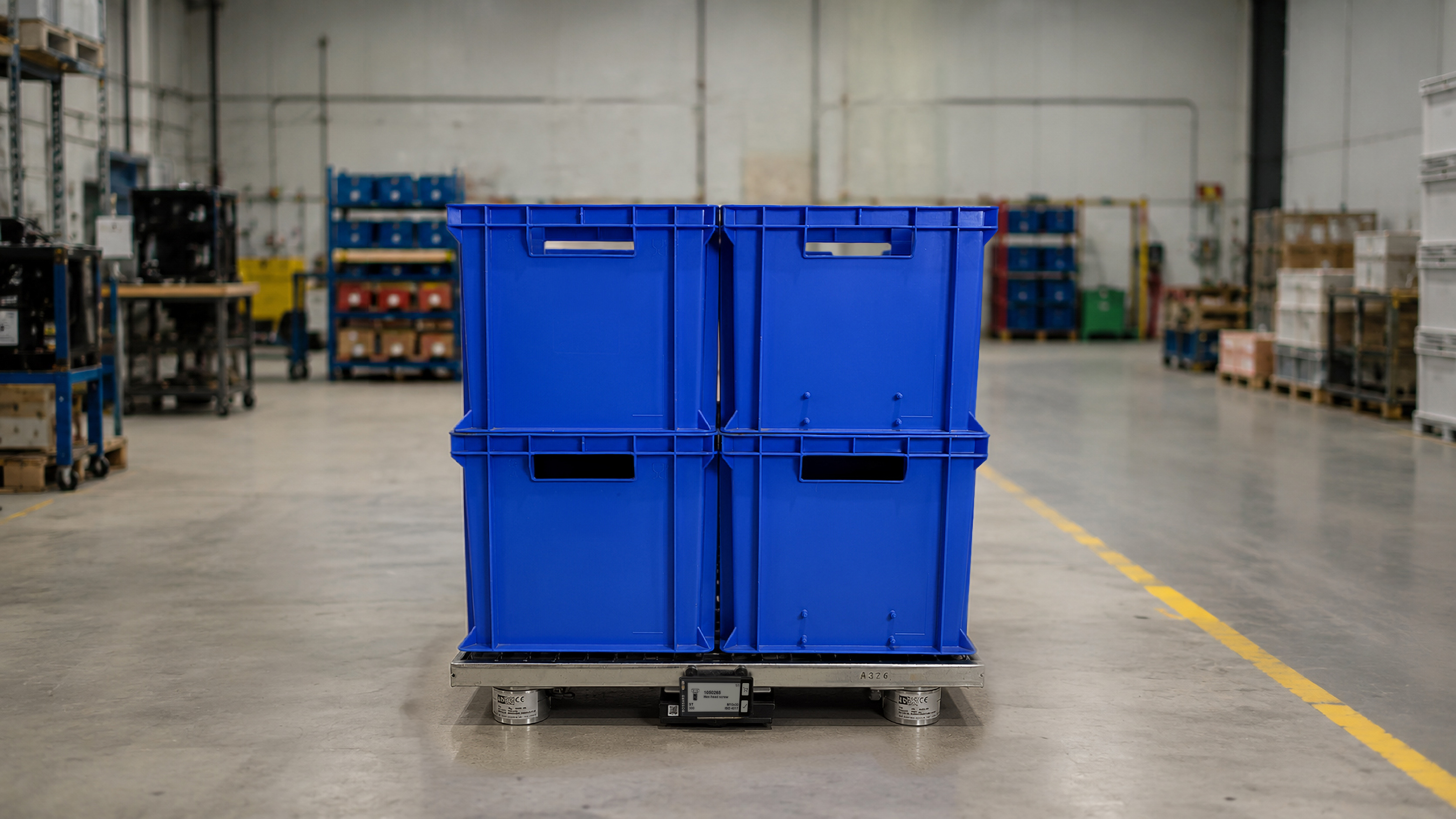 Blue plastic storage bins stacked on a smart pallet system in a warehouse with shelving and yellow safety posts on the floor.