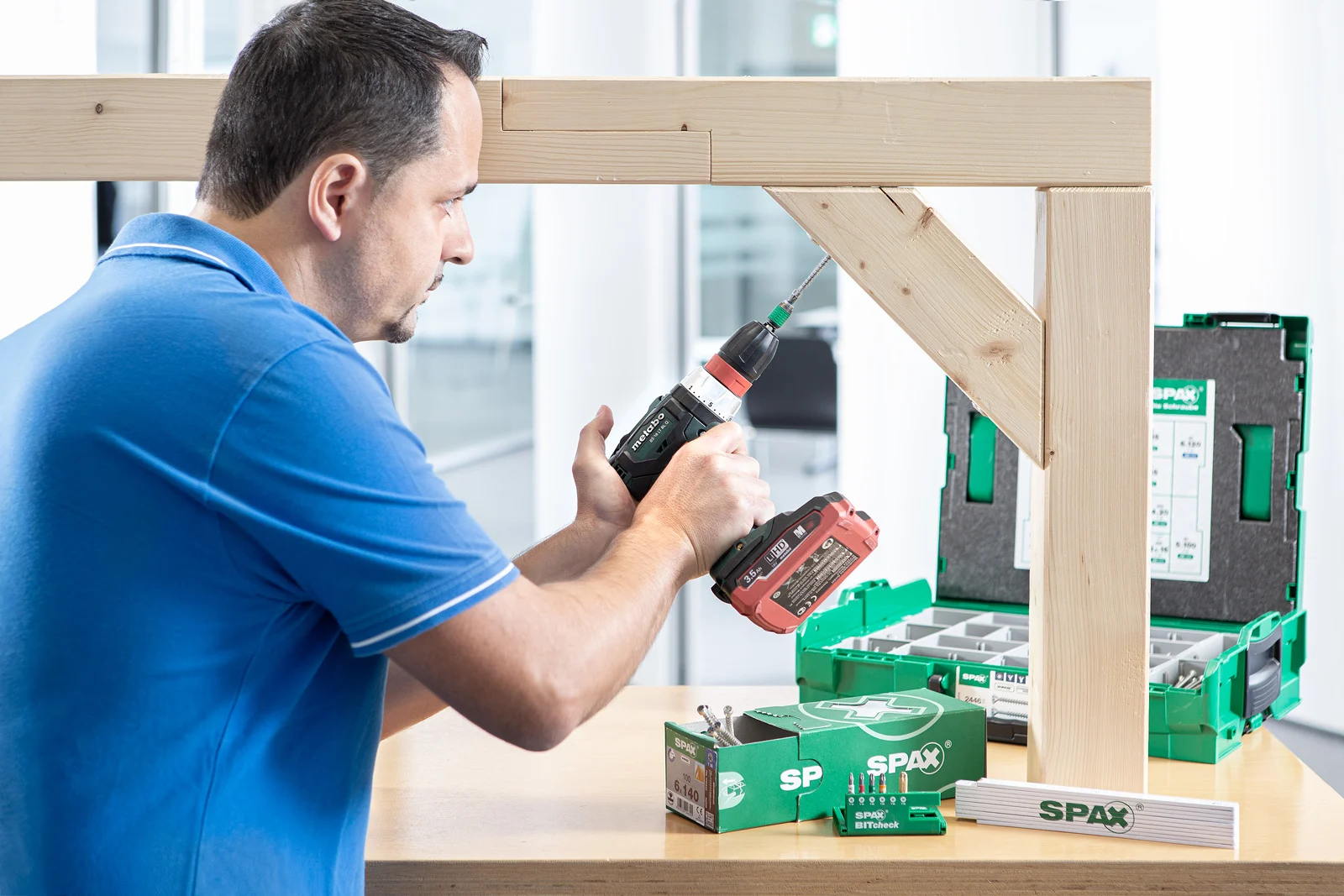 Ein Mann in blauem Polo verwendet eine Bohrmaschine, um einen Holzrahmen zu bauen.