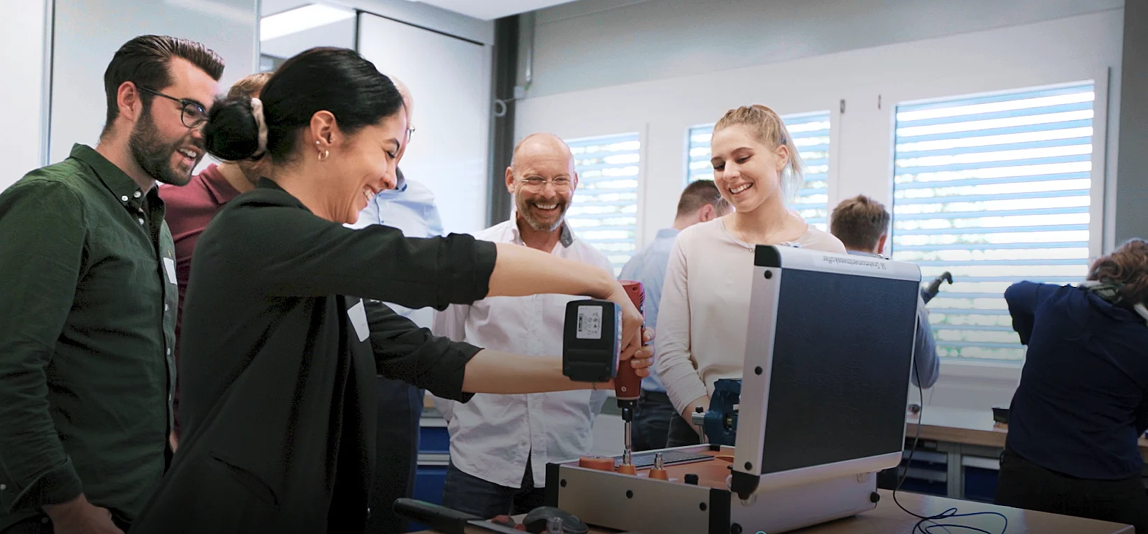 
A group of people stands around a workbench, using tools and a cordless screwdriver on the workbench.