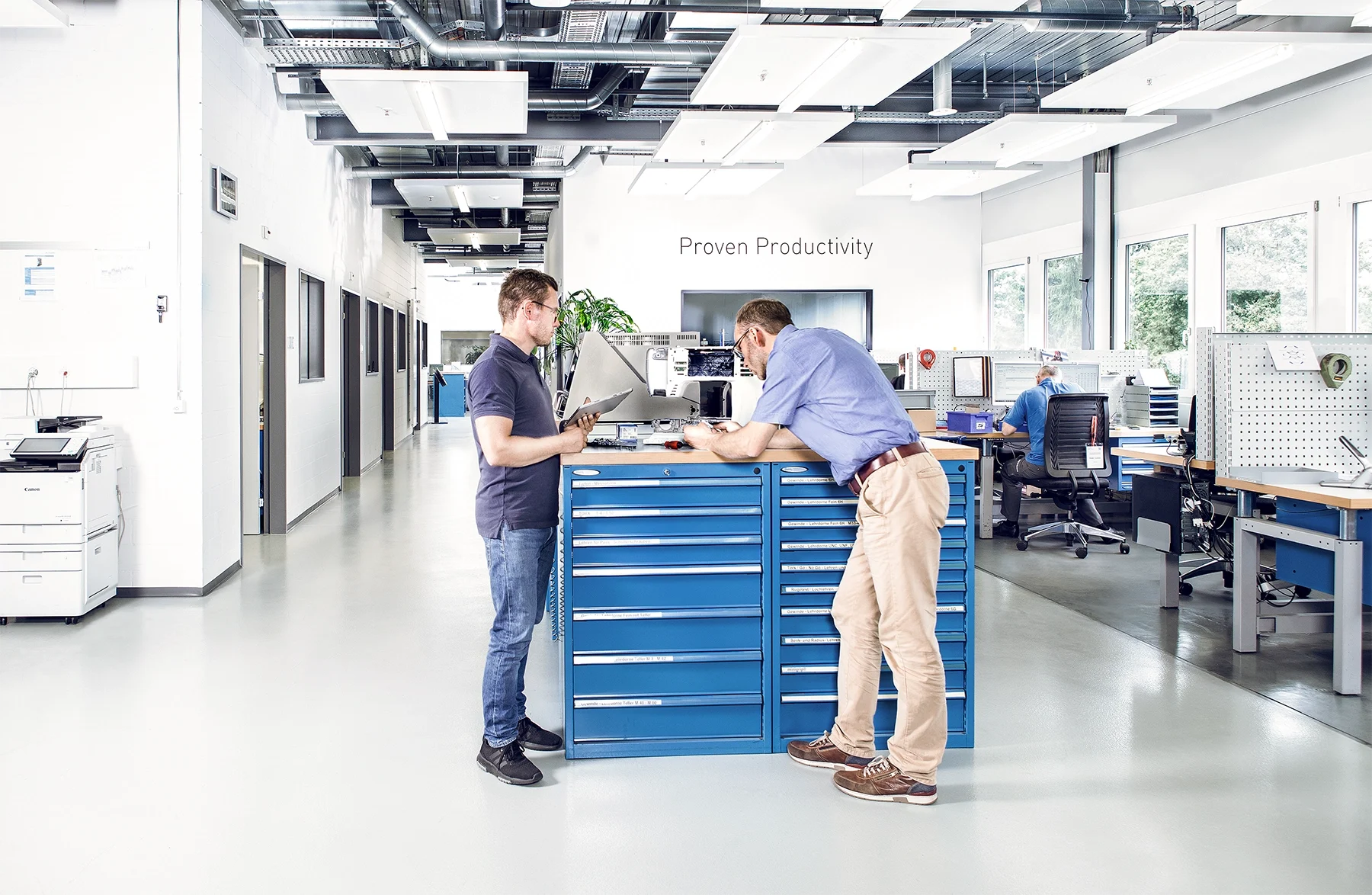 Two men in a test lab examine a sewing machine on a desk in an industrial lab with additional workstations in the background. Key visual for Expert Test Services.