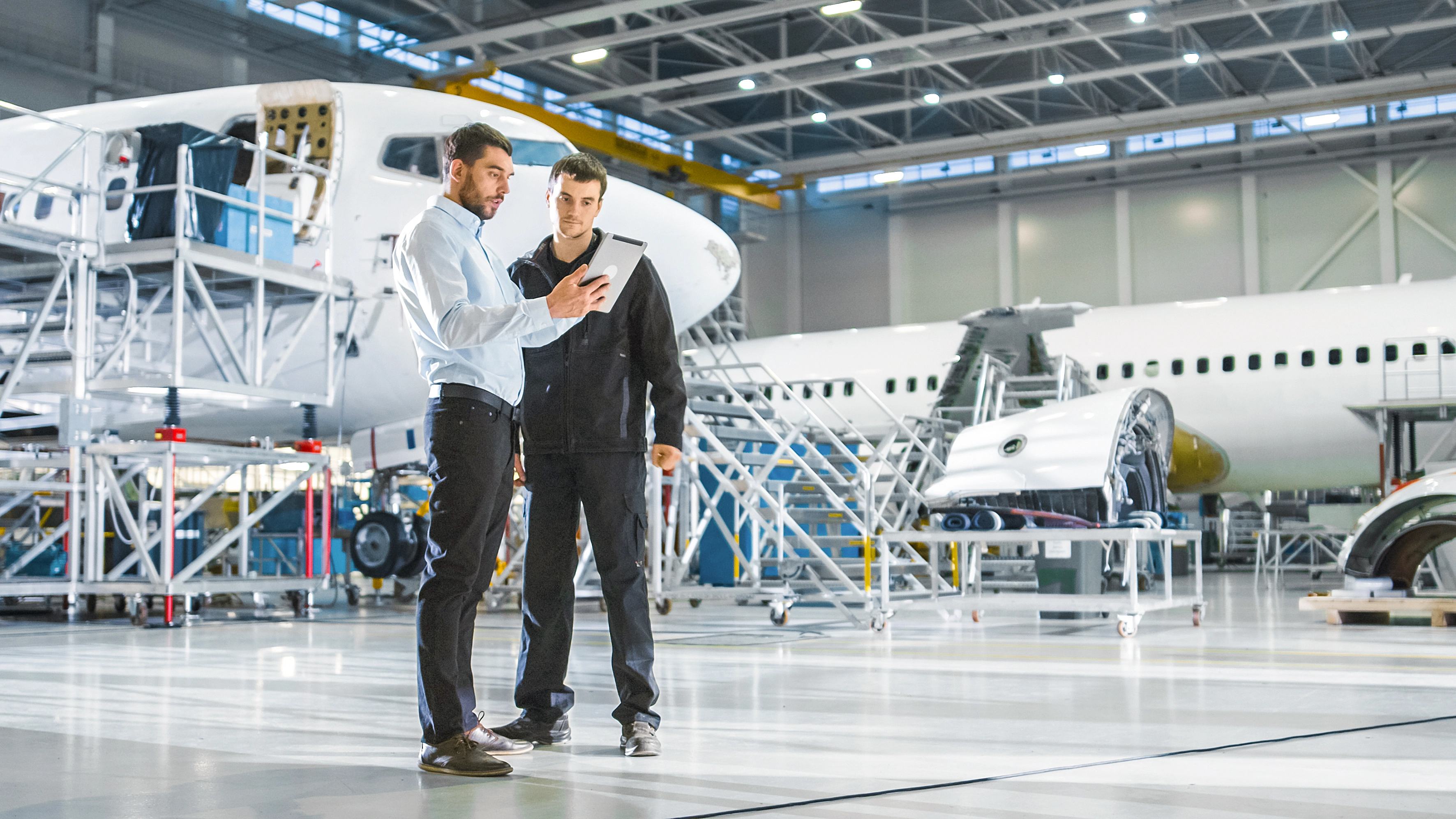 I en flyfabrik fokuserer to mænd på en tablet og analyserer information omgivet af flykomponenter.