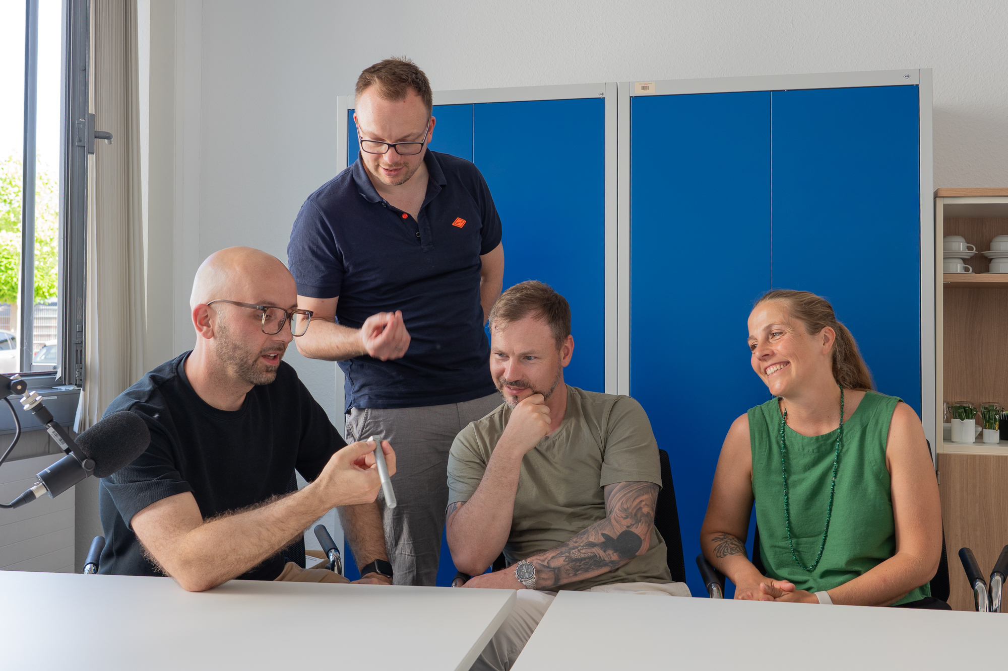 Vier Personen im Gespräch während einer Podcast-Aufnahme zum Thema DIN- und Normteile. Ein Teilnehmer hält ein Verbindungselement in der Hand, während ein Mikrofon im Vordergrund steht.