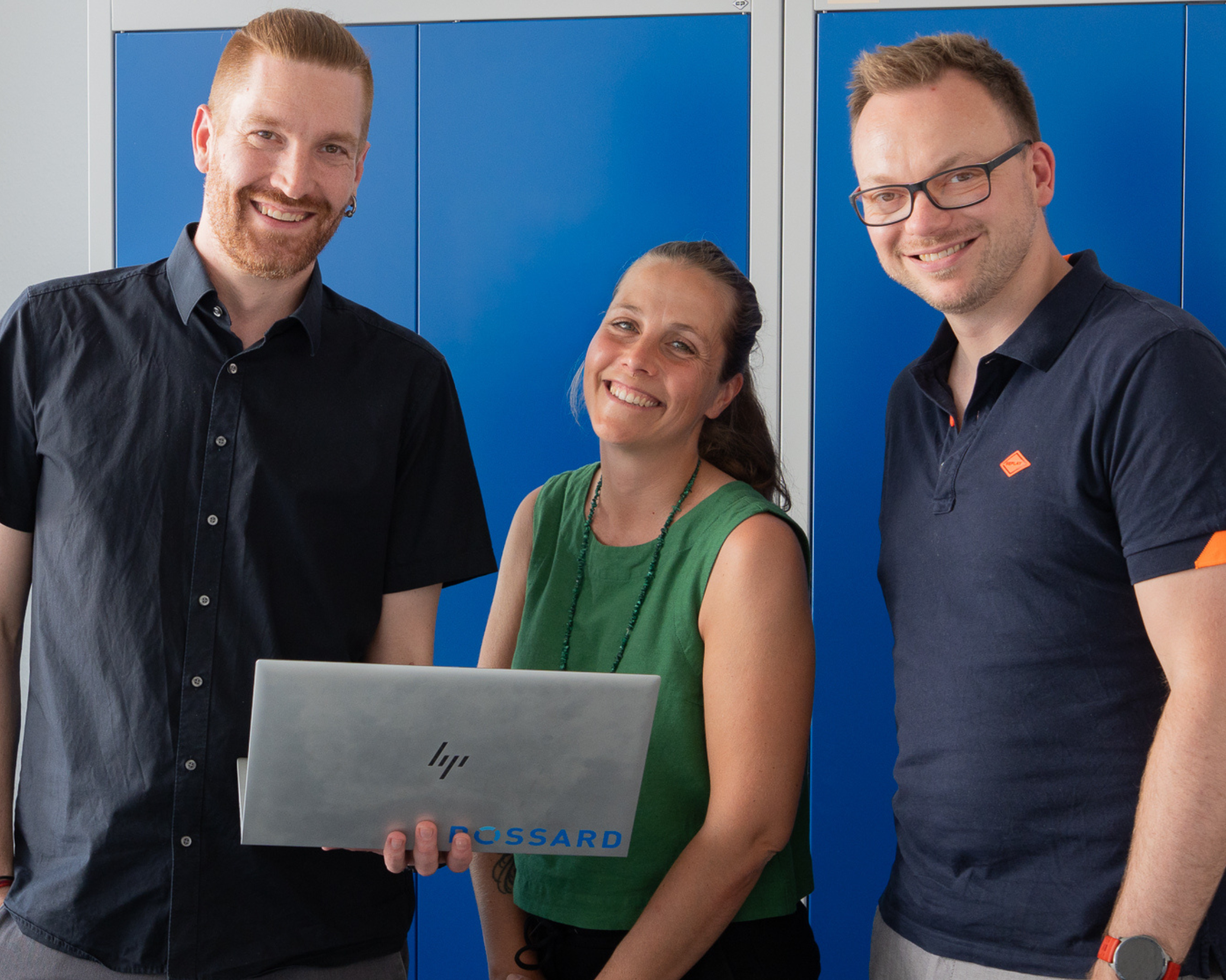 Matthias Schorer, Maja Matzke und Tobias Engelberg stehen lächelnd vor blauen Schränken; Maja Matzke hält einen geöffneten Bossard Laptop.