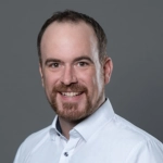 Portrait von Alex Dobler mit Bart, freundliches Lächeln, weißes Hemd, grauer Hintergrund.