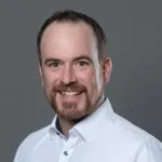 Portrait von Alex Dobler mit Bart, freundliches Lächeln, weißes Hemd, grauer Hintergrund.