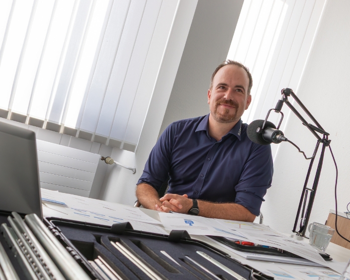 Alexander Dobler mit Mikrofon am Podcast-Tisch, umgeben von Teleskopschienen und Unterlagen, lächelnd vor dem Mikrofon.