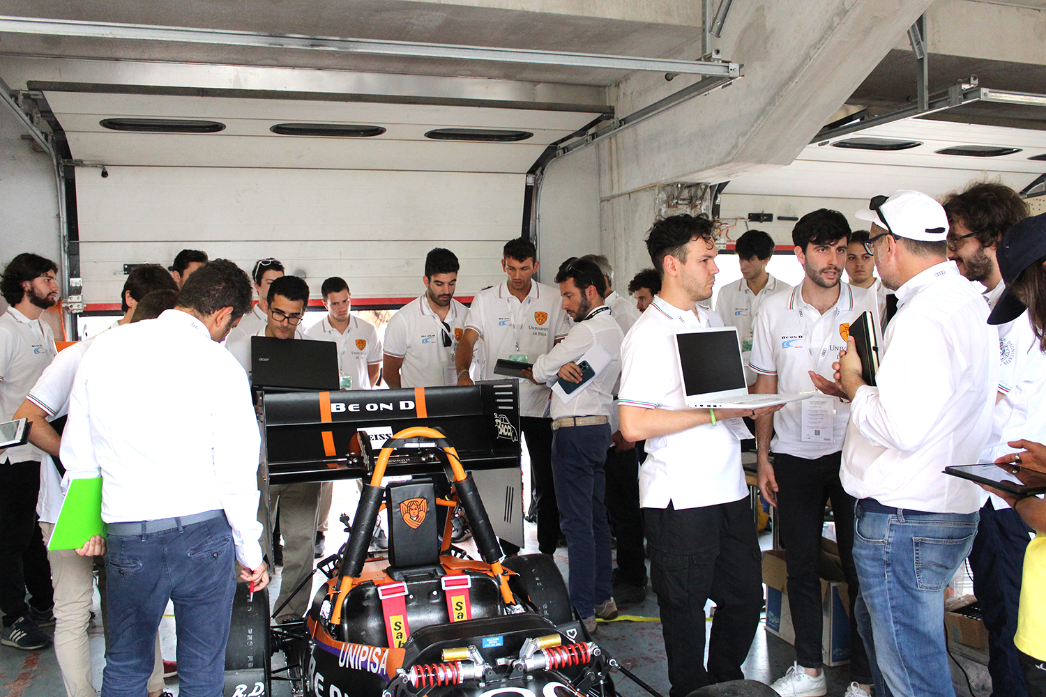 A group of people in white shirts gather around a race car in a garage, holding laptops and discussing.