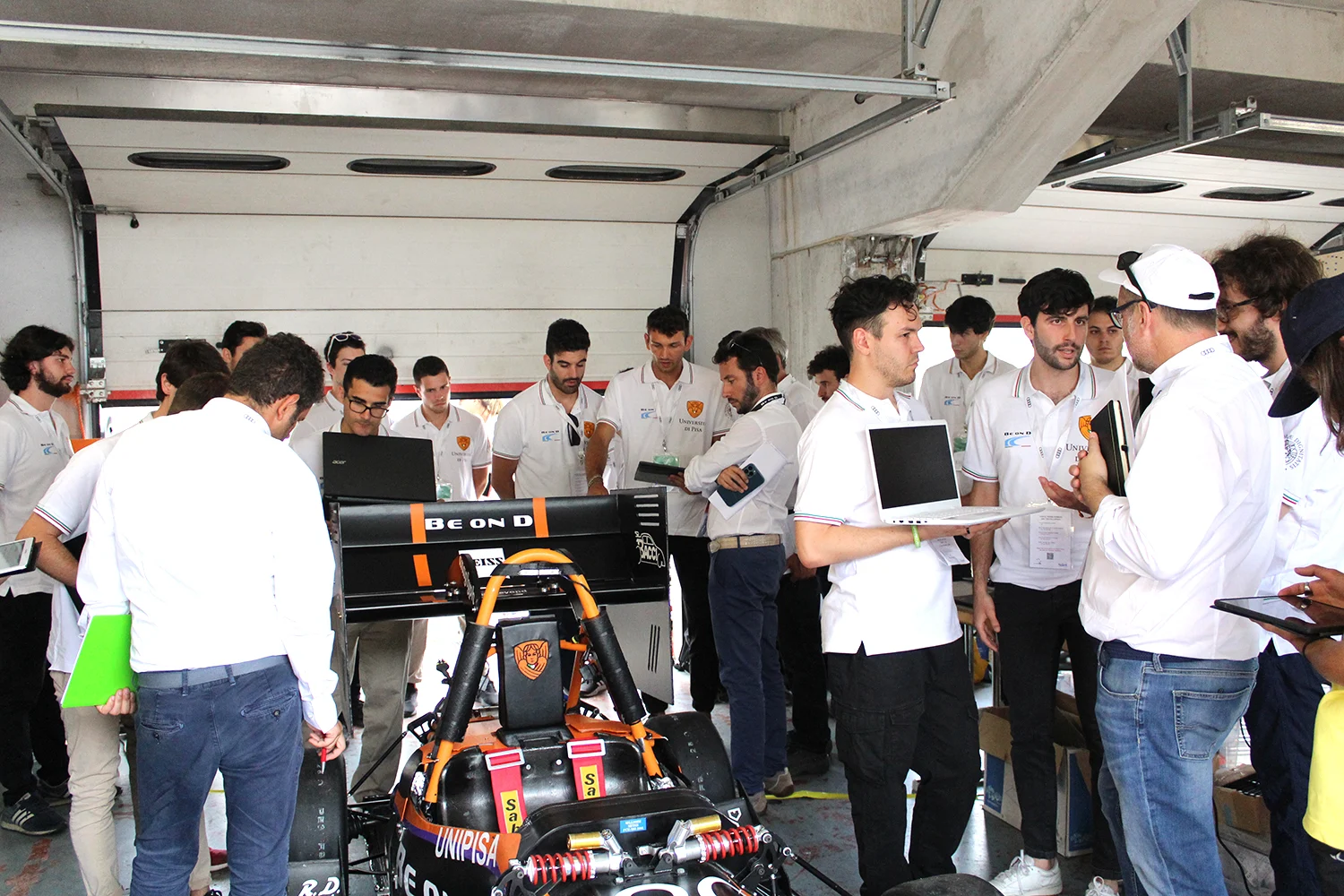 Un gruppo di persone in camicie bianche si riunisce attorno a un'auto da corsa in un garage, con laptop in mano e discutendo.