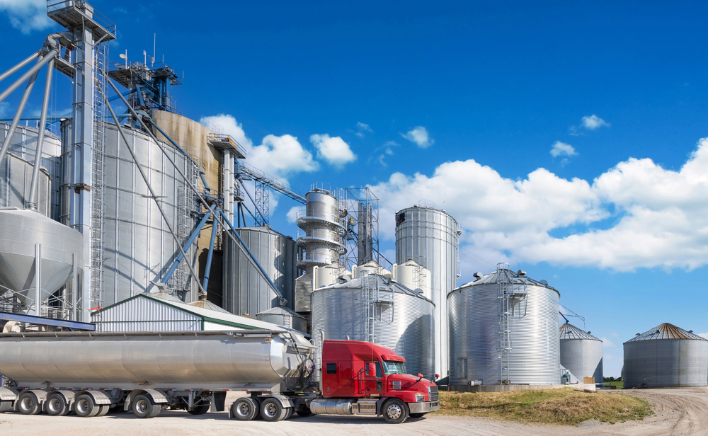 Red semi truck parked in front of large grain silos with open blue sky in the background