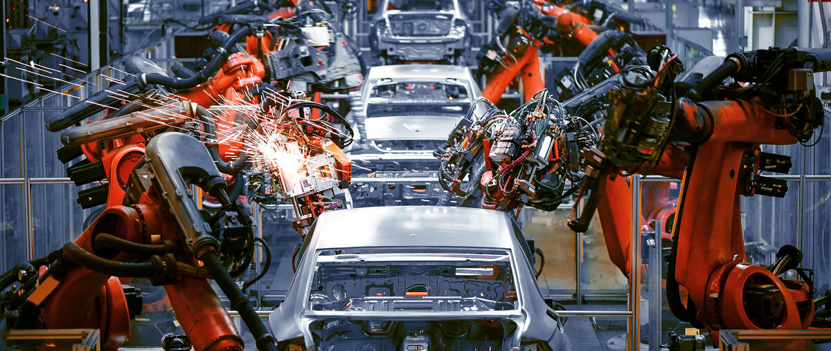 Orange and black robotic arms welding white car bodies on an automotive assembly line with sparks flying from welding points.