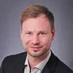 Portrait von Denis Heinrich, leichtes Lächeln, blauer Hintergrund.