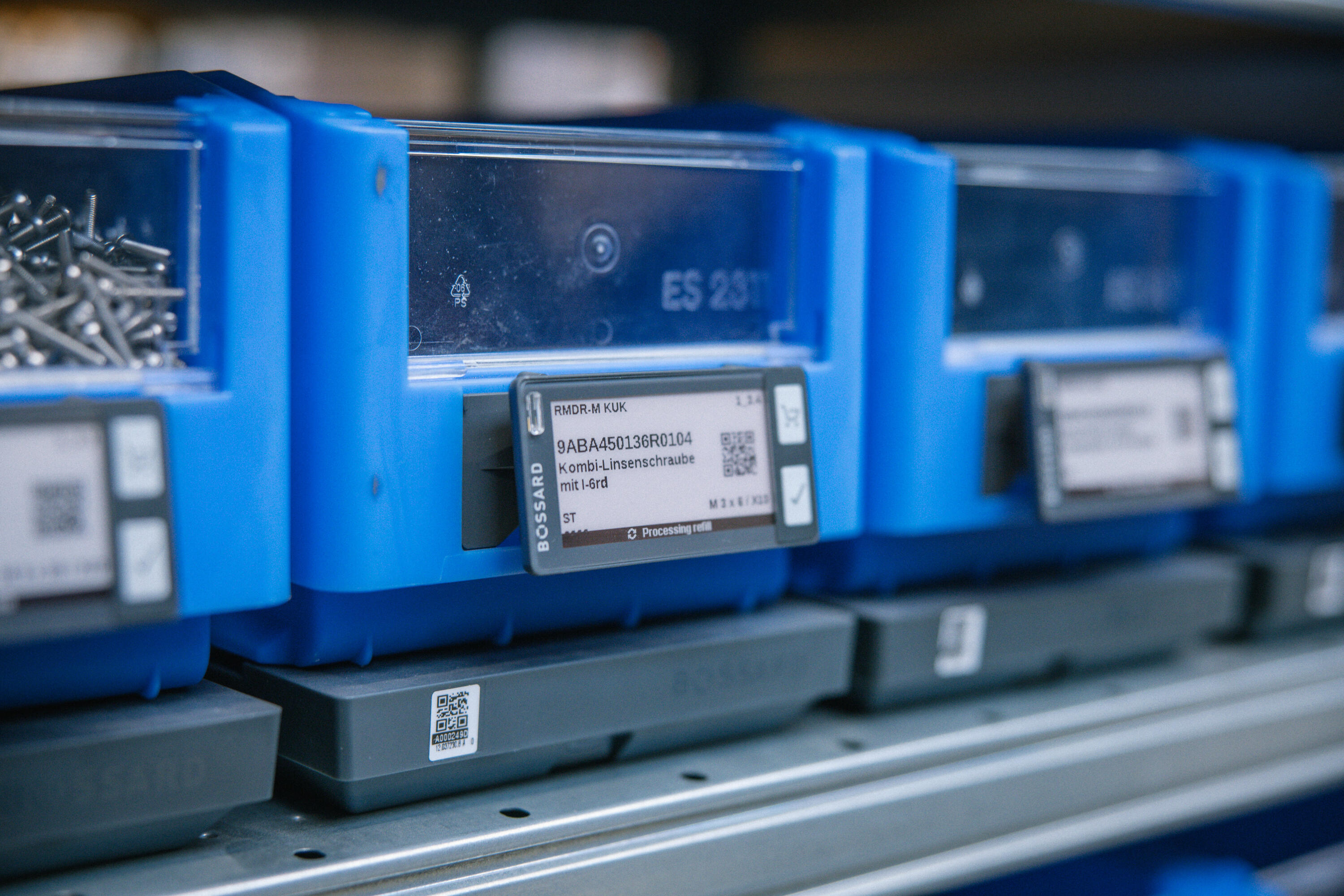 Blue plastic storage bins with clear fronts and digital labels displaying part numbers, arranged on a shelf system.
