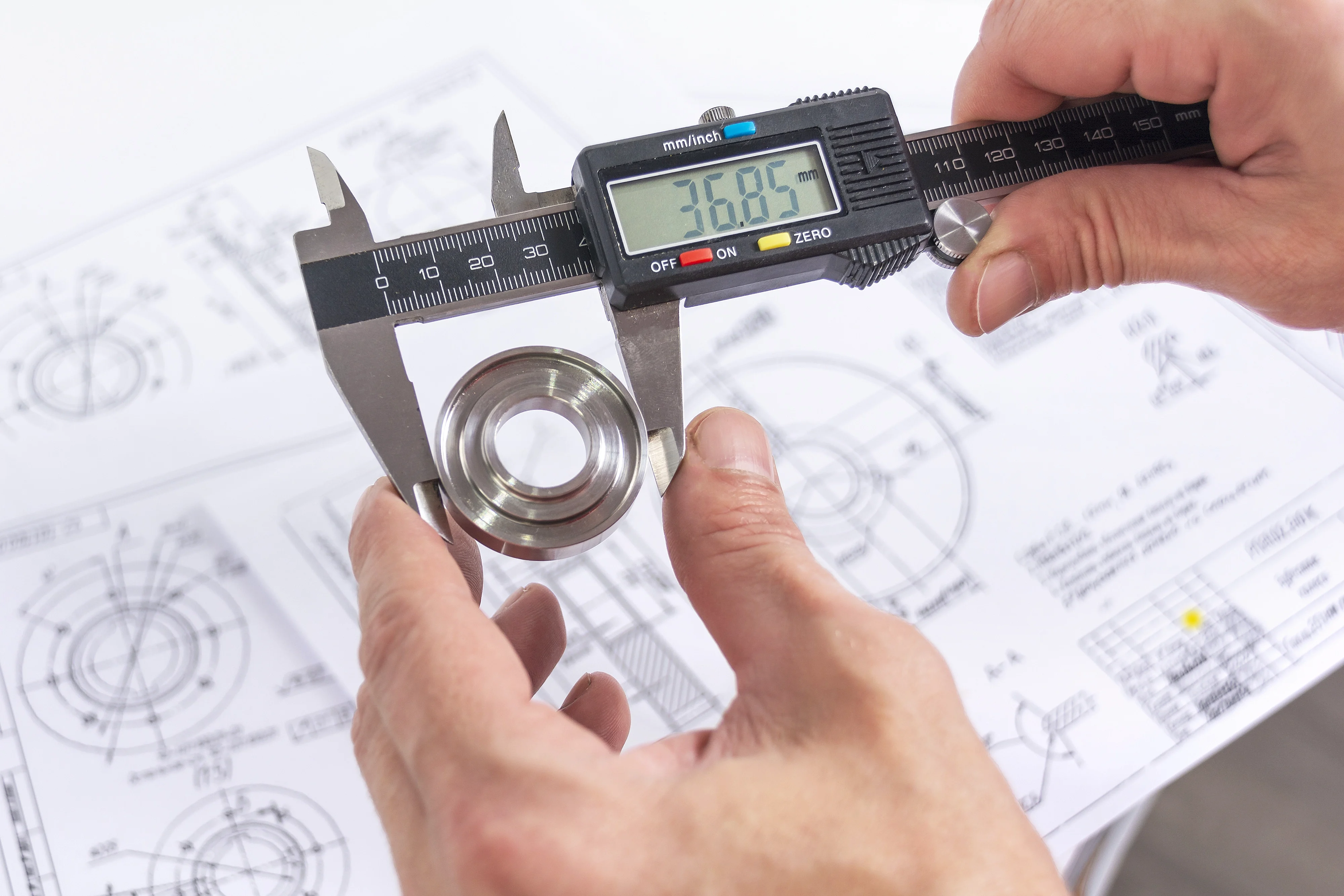 A pair of hands gripping a caliper to measure a metal bearing above technical drawings and specifications.