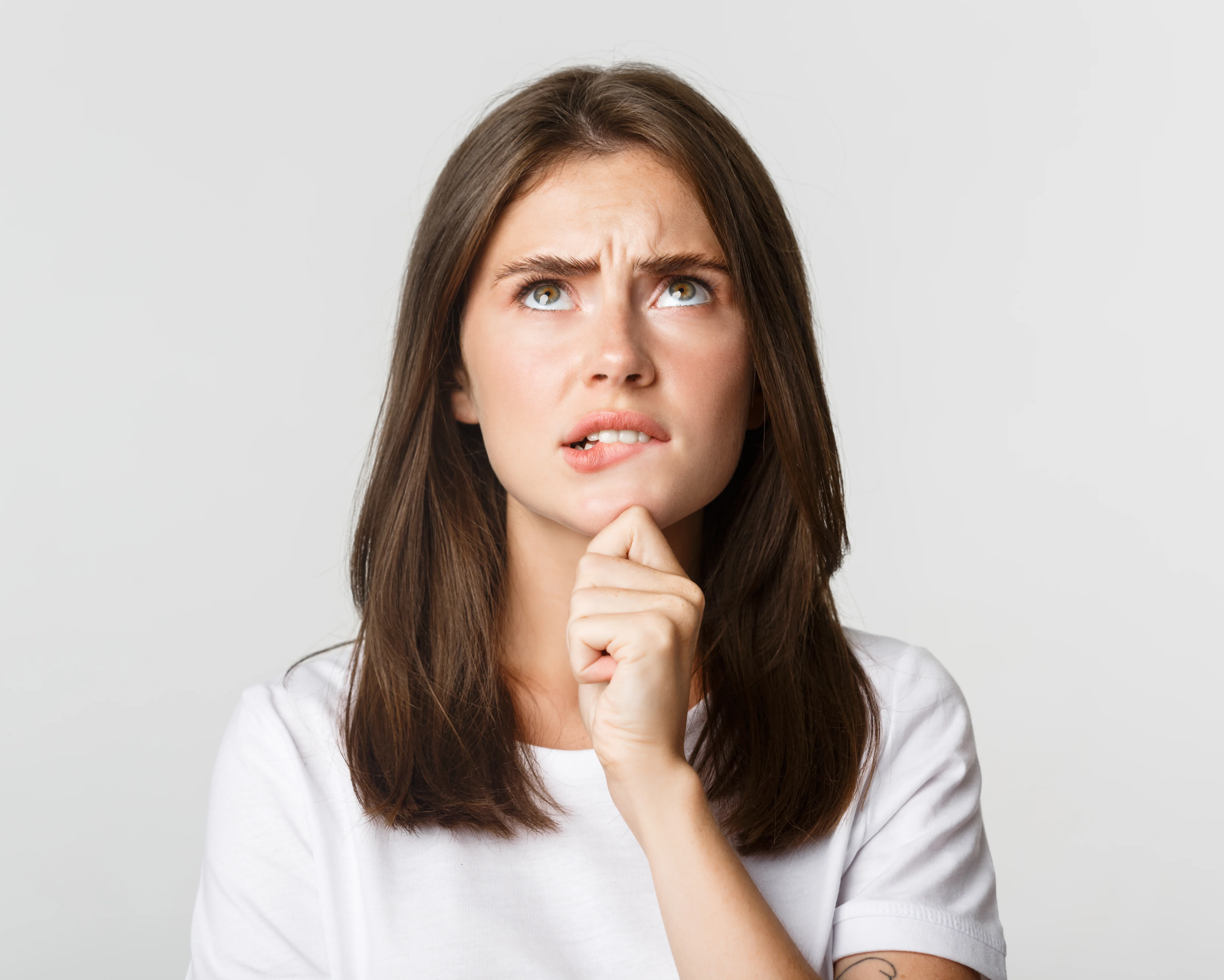Frau mit langen braunen Haaren in einem weißen Hemd, die nachdenklich nach oben schaut, Hand am Kinn, vor einem schlichten grauen Hintergrund.