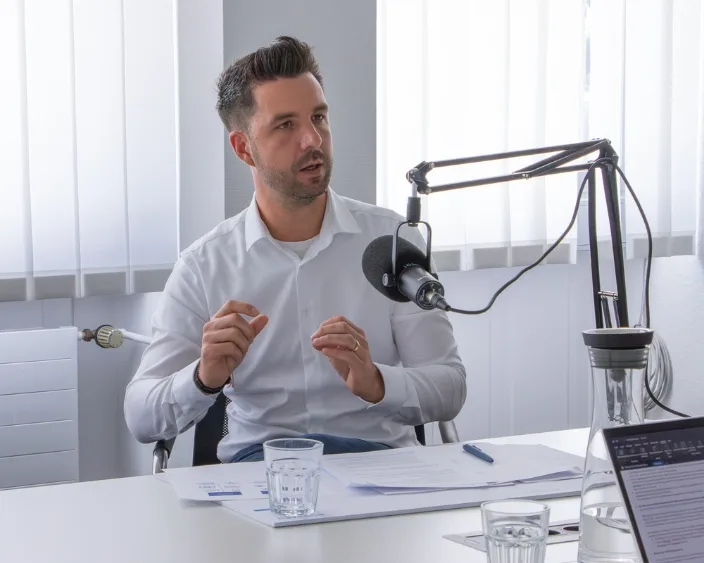 Manuel Ohmayer im weißen Hemd beim Podcast-Interview, sitzend am Tisch mit Mikrofon und Unterlagen.