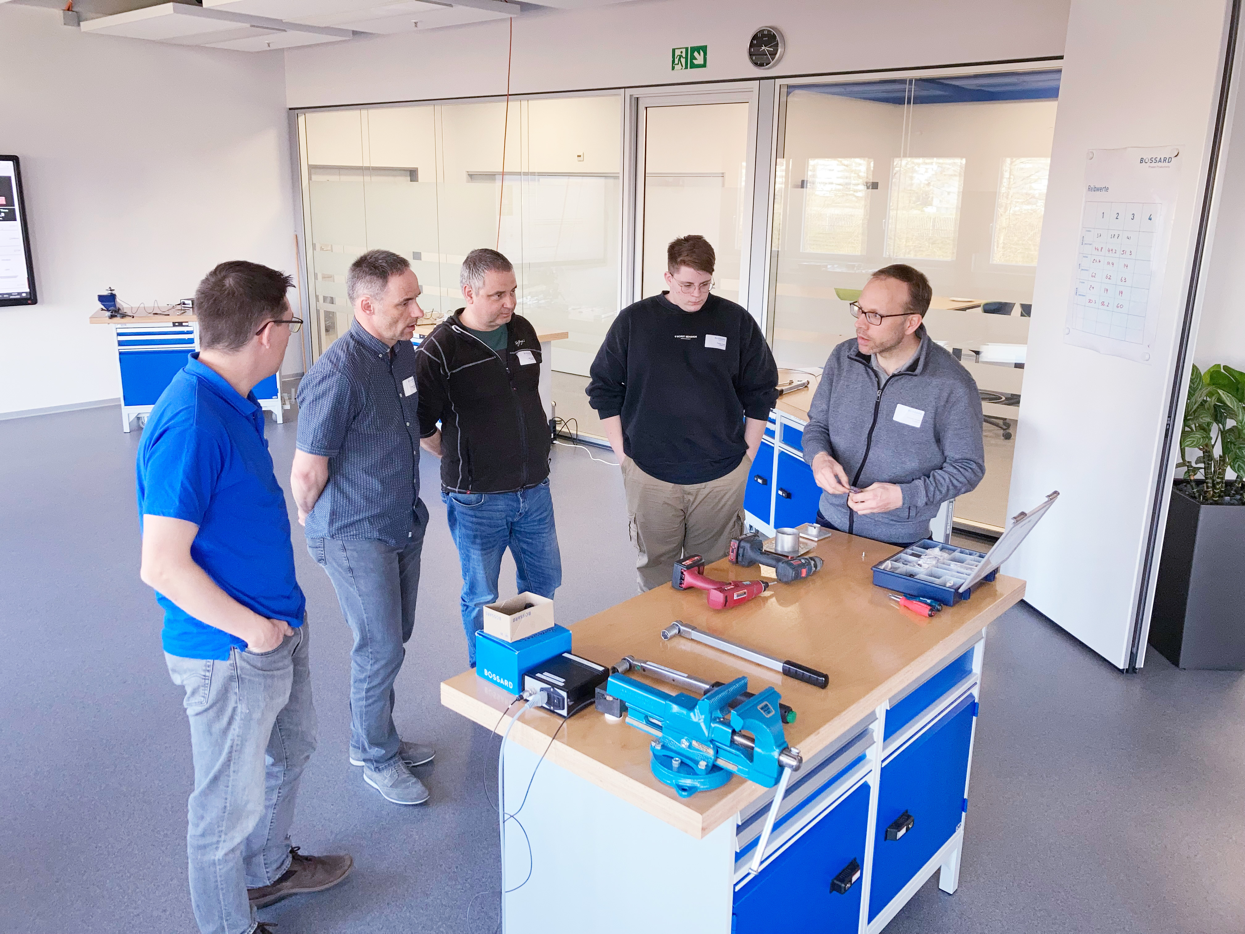 A group of men stand around a workbench with tools and discuss together.