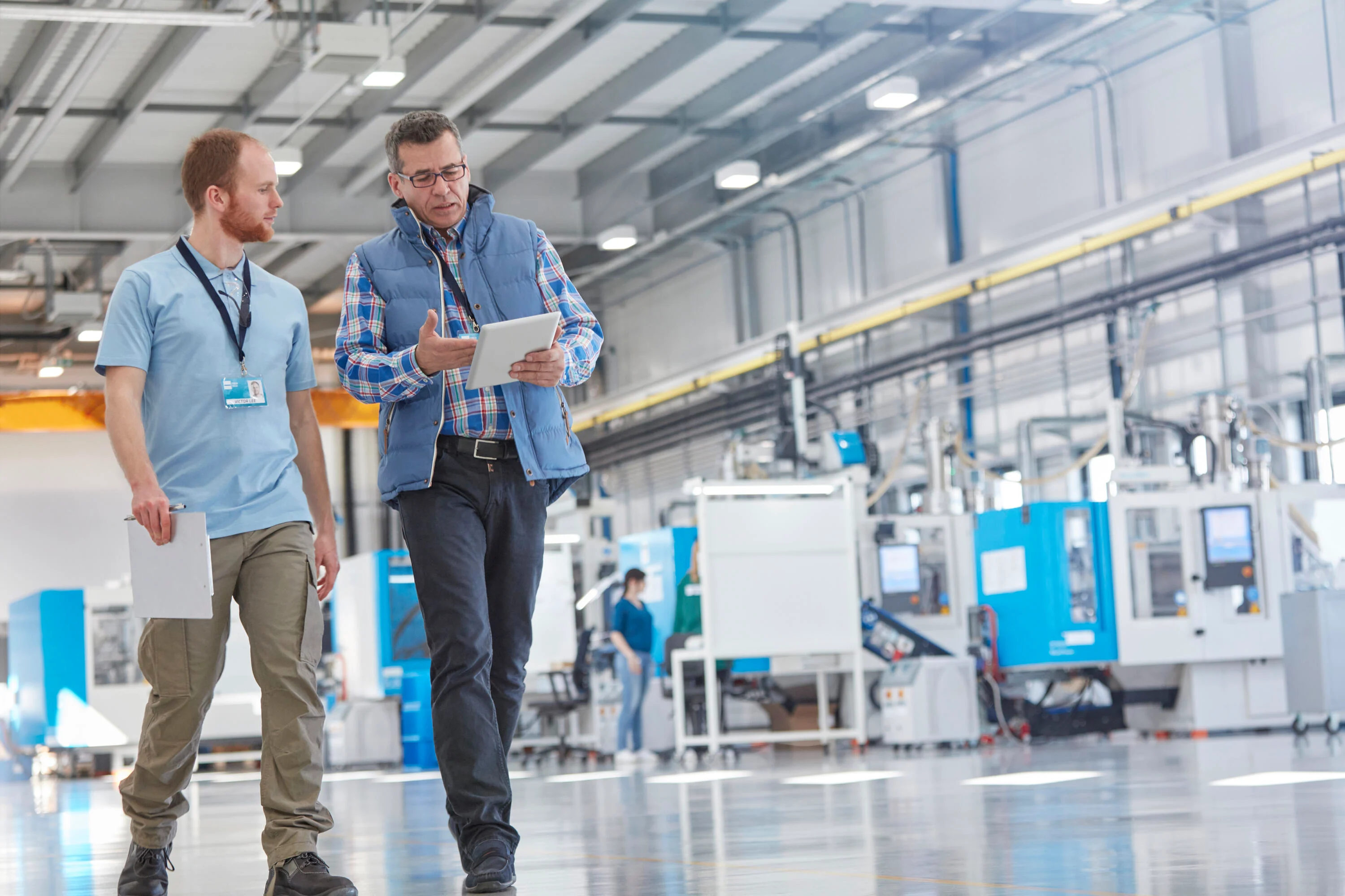 Two men walking through a customer site's production line. One holds a tablet, explaining to the other. Background shows the production/assembly line. Key visual for Expert Walk only.