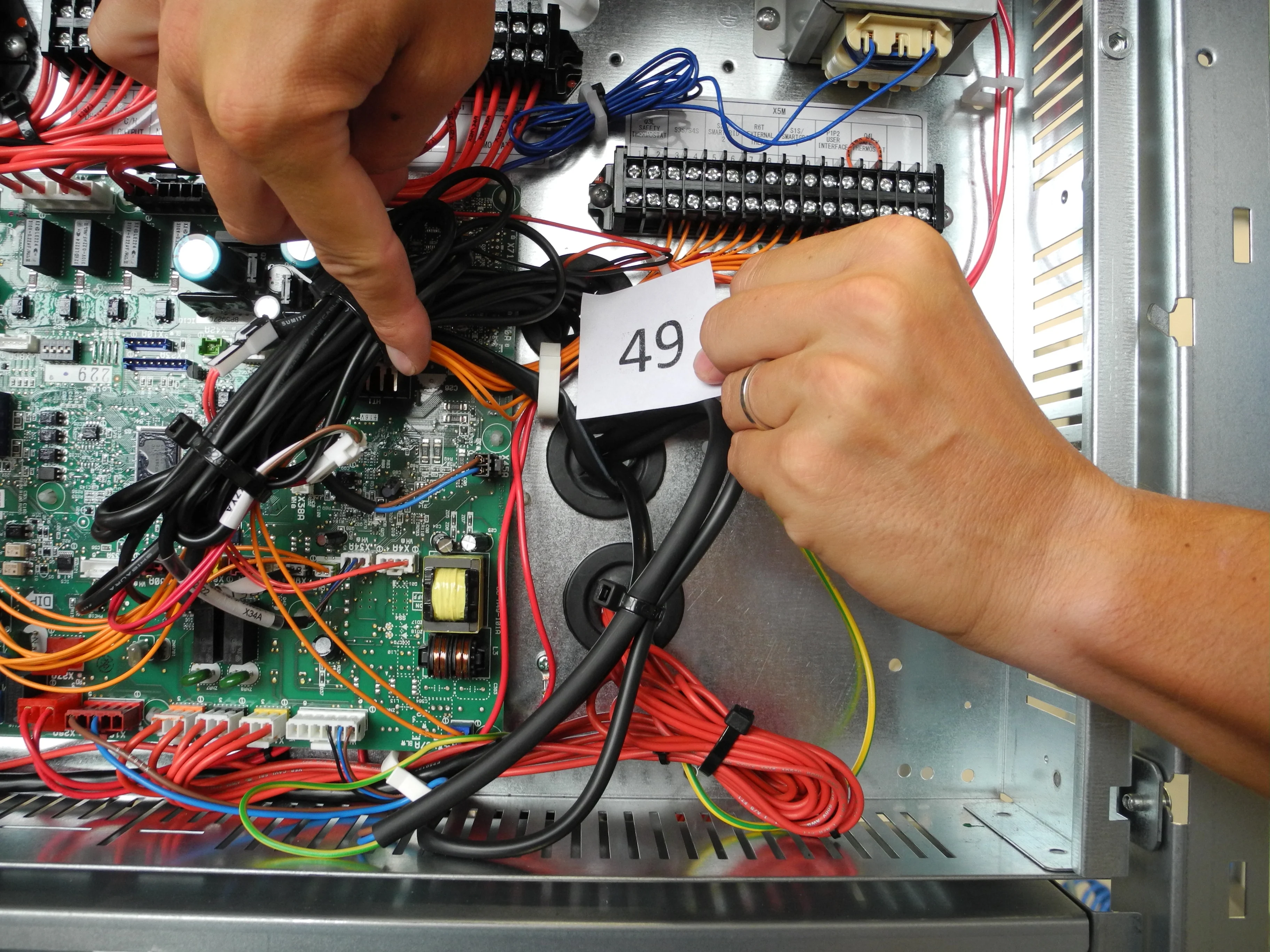 Ruce technika označující drát číslo 49 uvnitř elektrického rozvaděče s deskami plošných spojů a barevným zapojením během Expert Teardown.