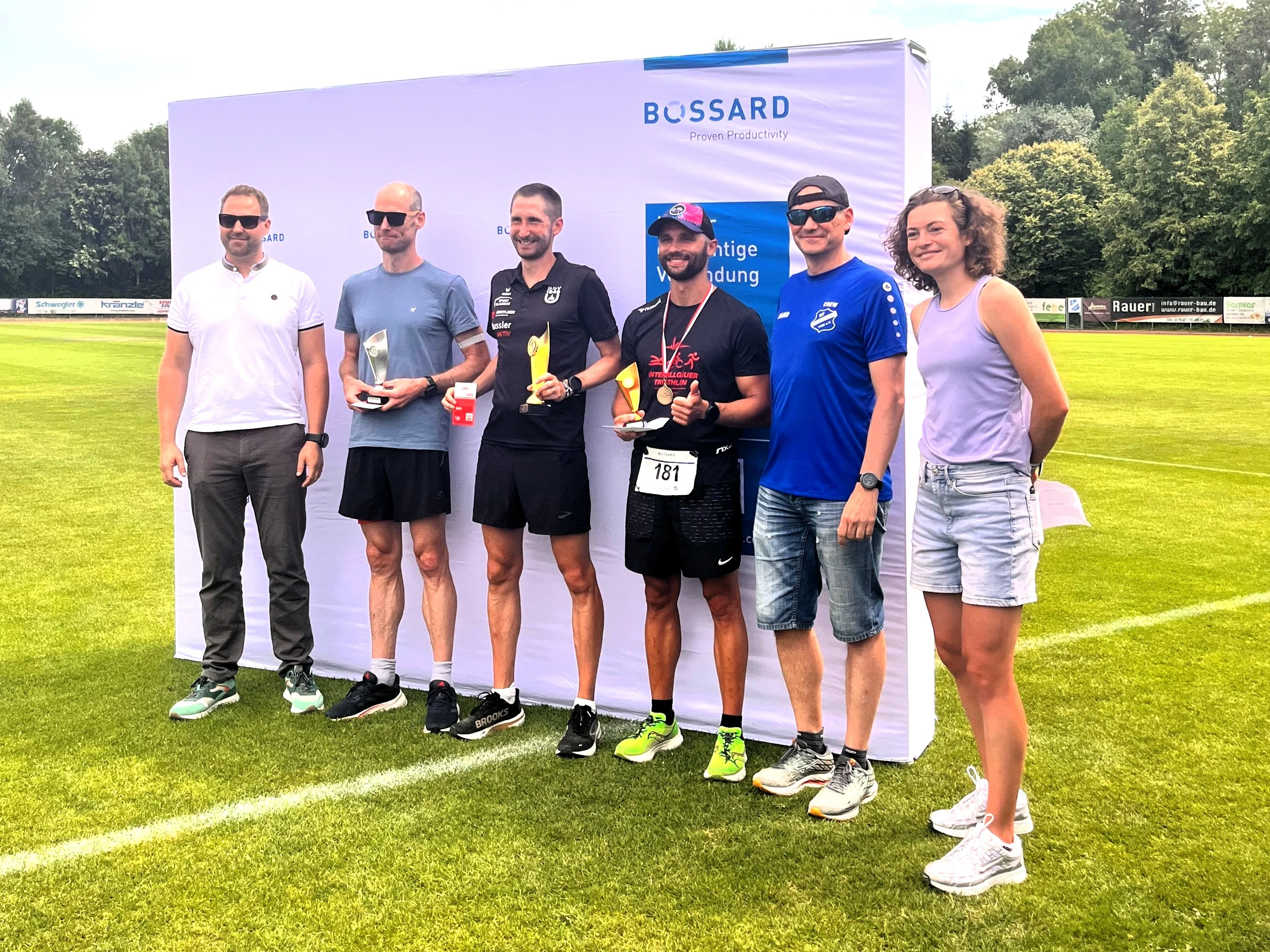 Gruppenfoto von sechs Personen inklusive Lauf-Siegern mit Pokalen vor einer Bossard-Wand auf einem Sportplatz.