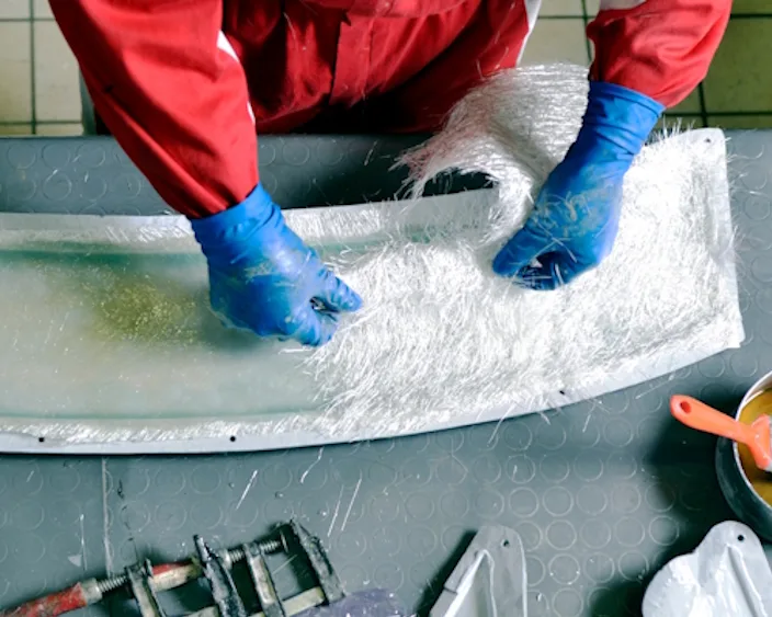 Person in roten Arbeitskleidung und blauen Handschuhen verarbeitet Glasfasermatten auf einer Form.