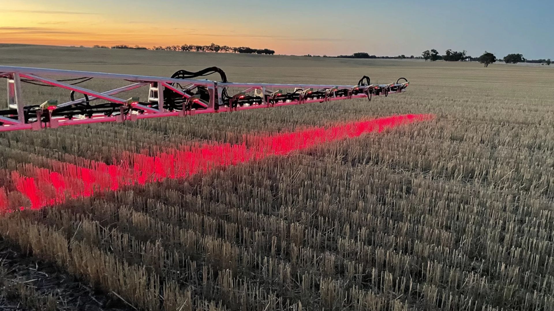 Rode landbouwspuitapparatuur die helder rood licht werpt over een geoogst stoppelveld bij zonsondergang met bomen aan de horizon.