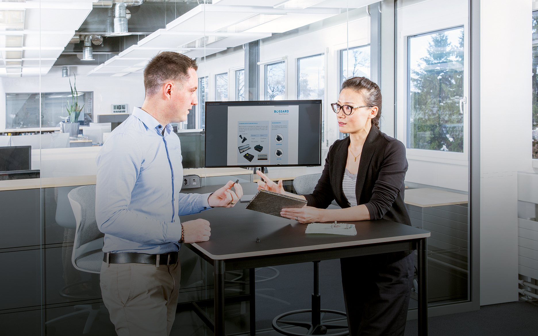 Two people at a desk in a modern office discuss fastening a composite material; one holds a sandwich panel, the other a fastener.