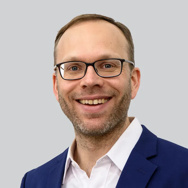 Ein Mann mit Brille und blauem Anzug lächelt freundlich in die Kamera.