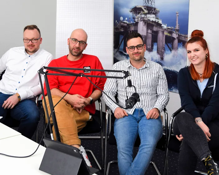 Drei Männer und eine Frau sitzen entspannt in einem Podcast-Studio mit Mikrofonen vor sich