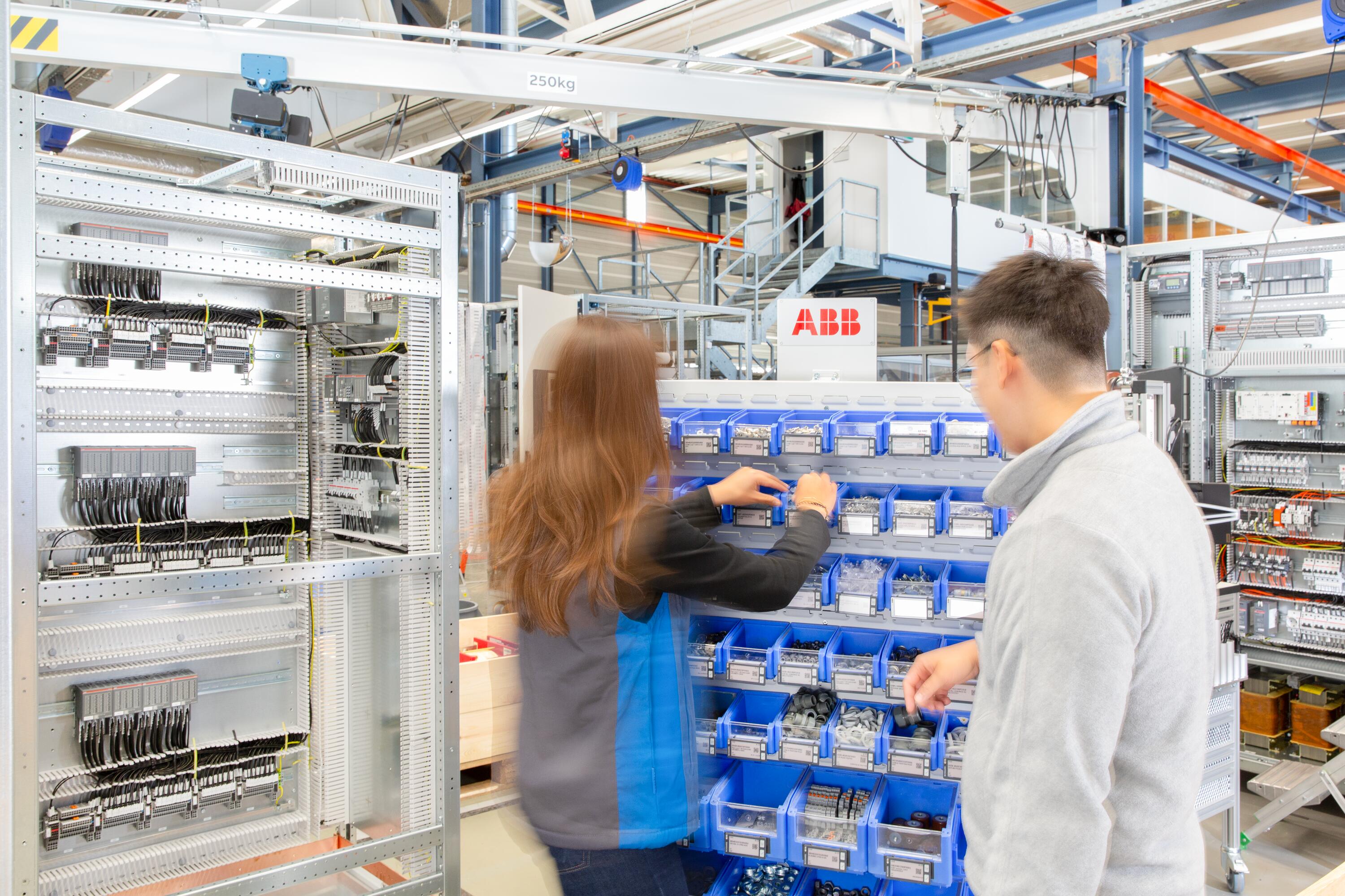 Two technicians in uniforms working at blue storage bins in an industrial facility with electrical panels.