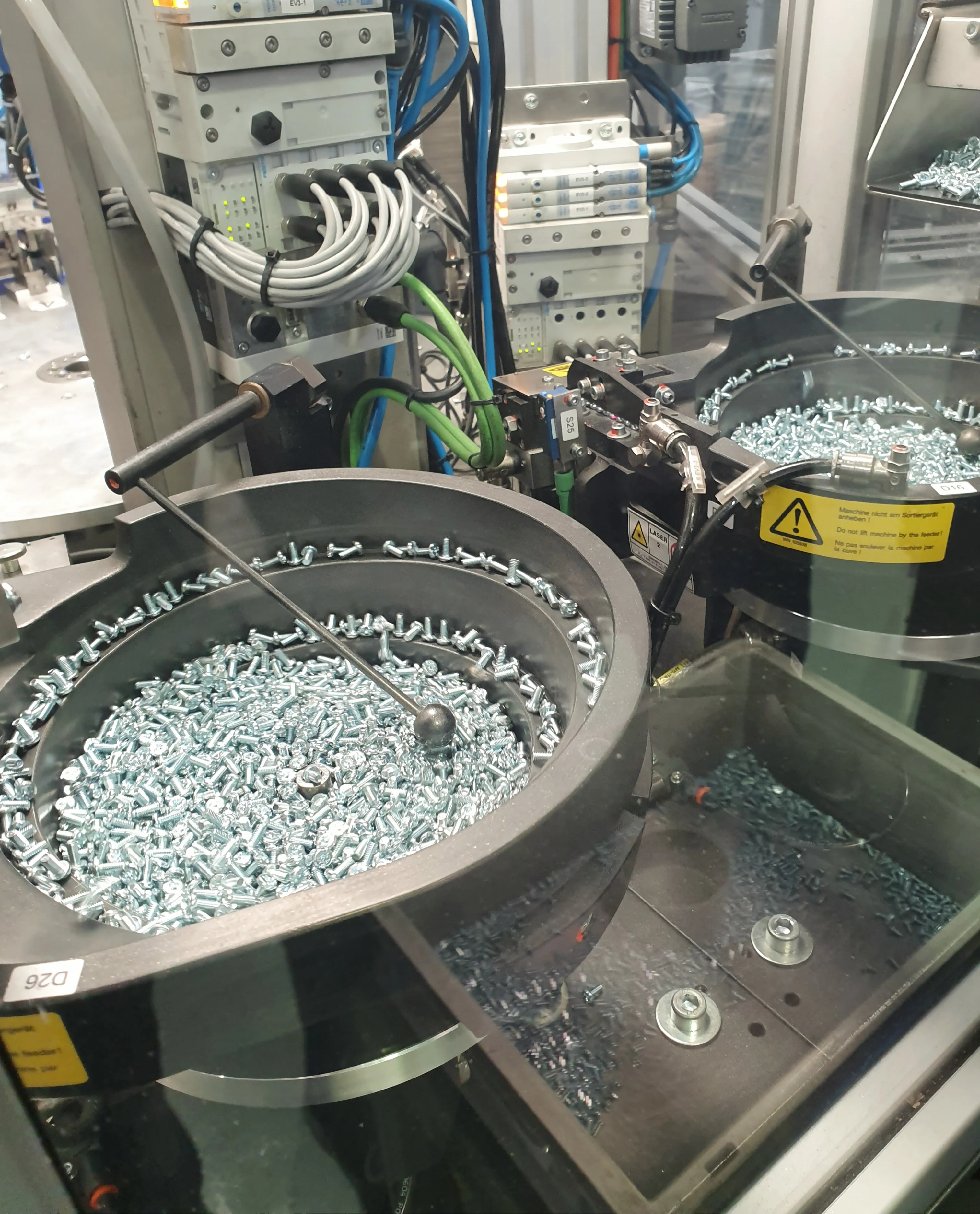 Máquina industrial con dos bandejas circulares llenas de tornillos plateados, rodeada de cables y paneles de control.