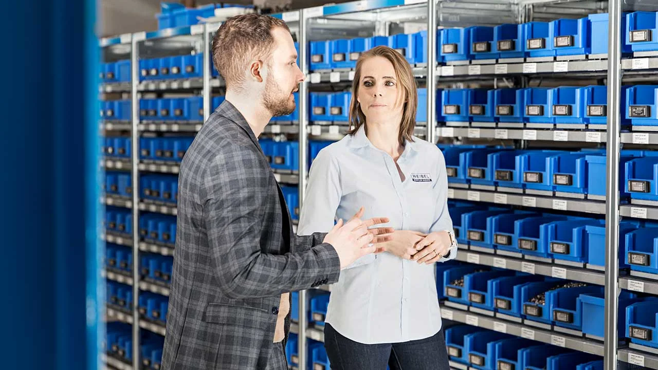 A man and a woman standing in front of a rack of blue Bossard Smartbins at Weibel