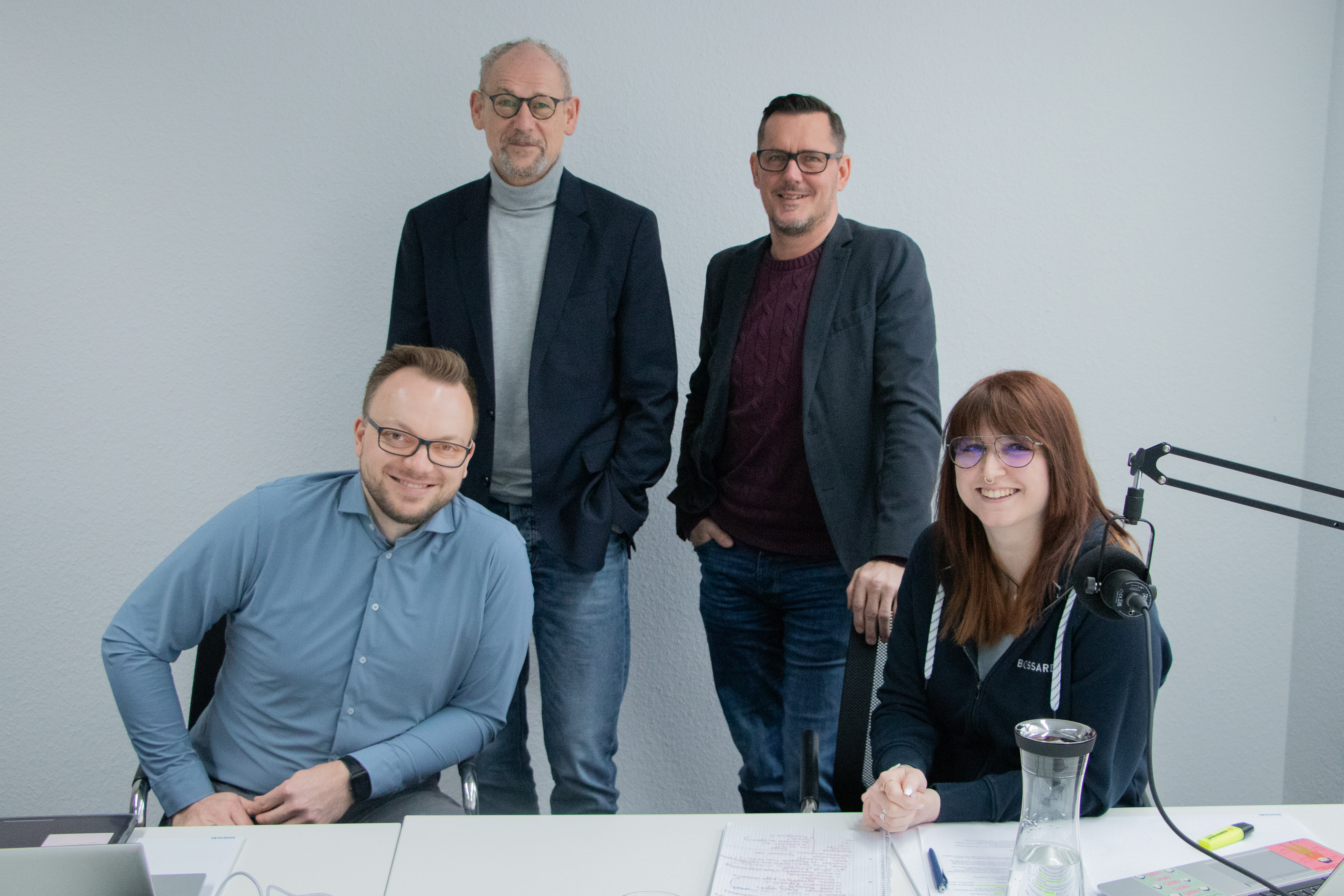 Vier Experten sitzen im Aufnahmestudio, weiße Wand im Hintergrund, Broadcast Mikrofone auf dem Tisch im Vordergrund