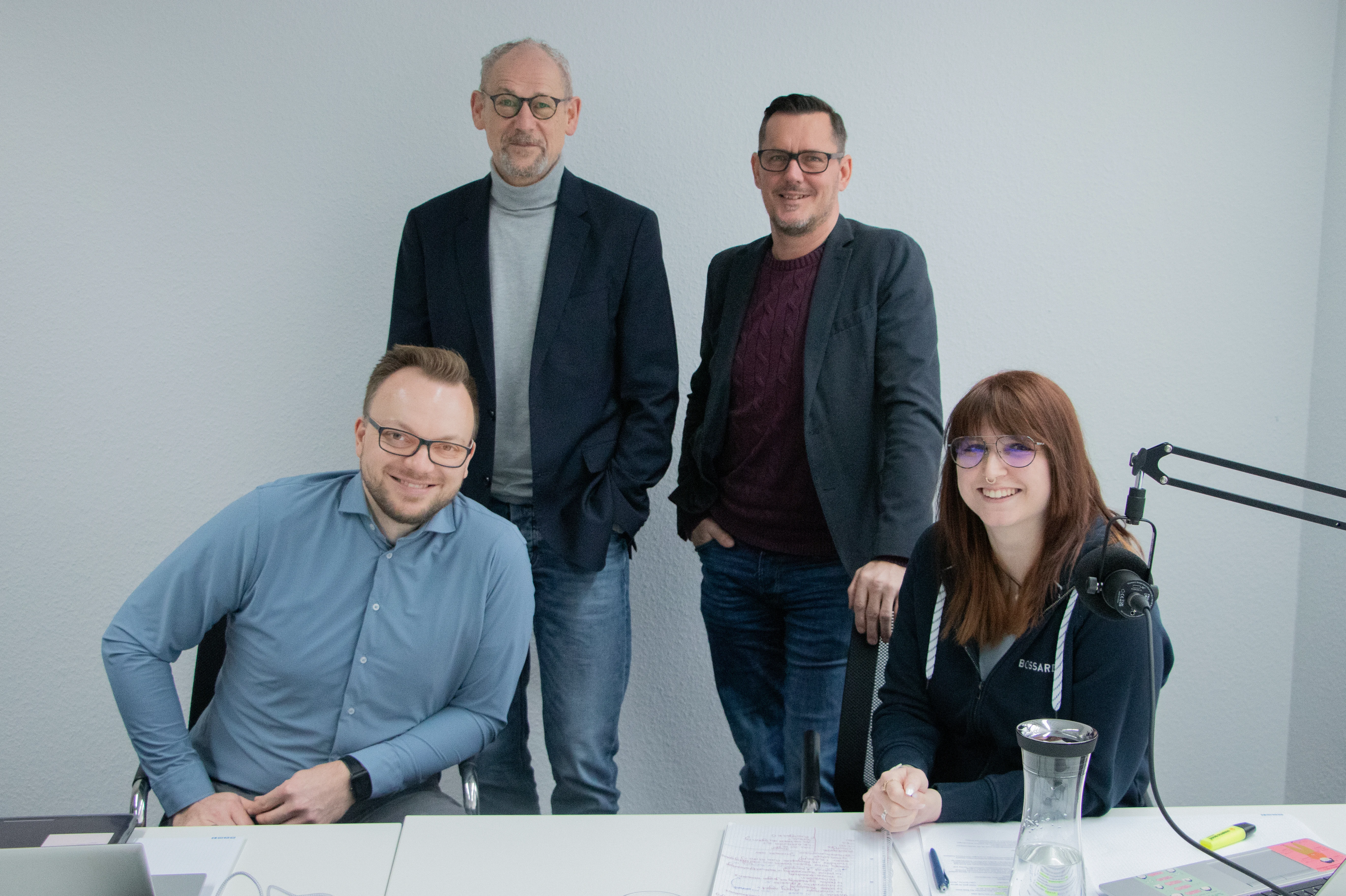 Vier Experten sitzen im Aufnahmestudio, weiße Wand im Hintergrund, Broadcast Mikrofone auf dem Tisch im Vordergrund