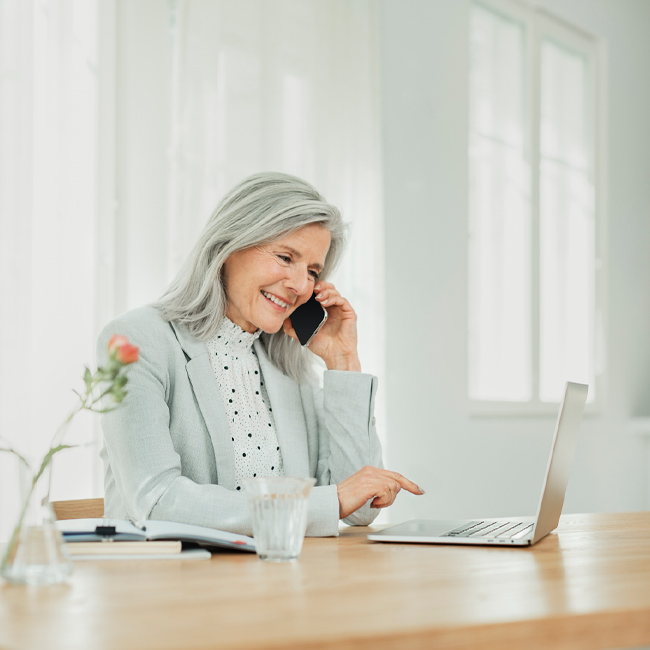 A woman sits at a table talking on the phone and looks at the laptop on the table.