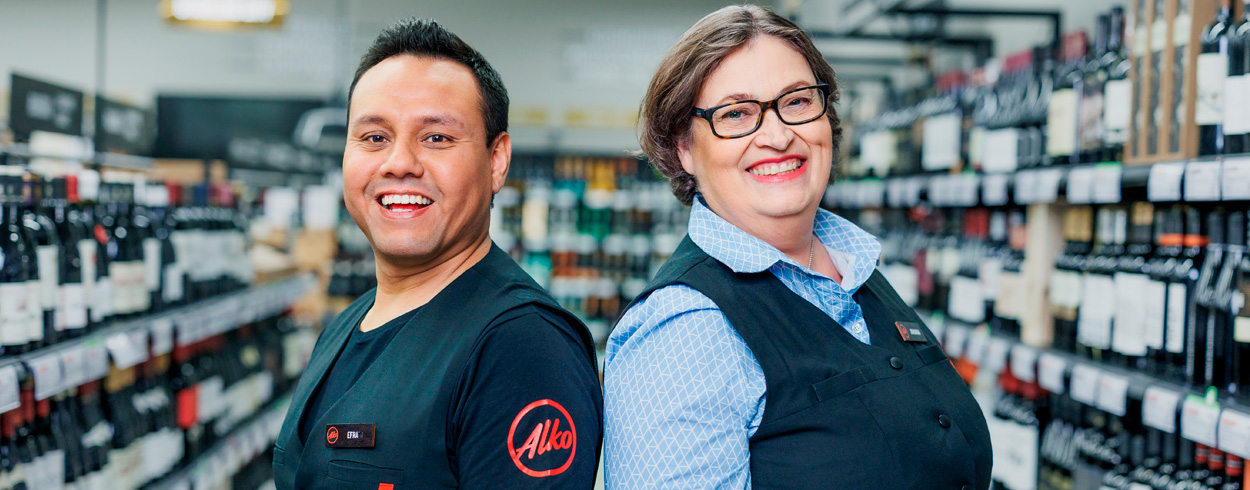 Kaksi hymyilevää myyjää selät vastakkain katsoo kameraan Alkon myymälässä hyllyjen välissä.