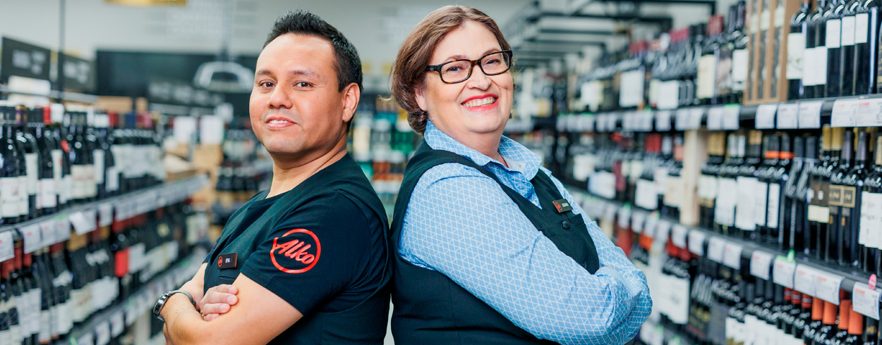 Kaksi hymyilevää myyjää selät vastakkain katsoo kameraan Alkon myymälässä hyllyjen välissä.