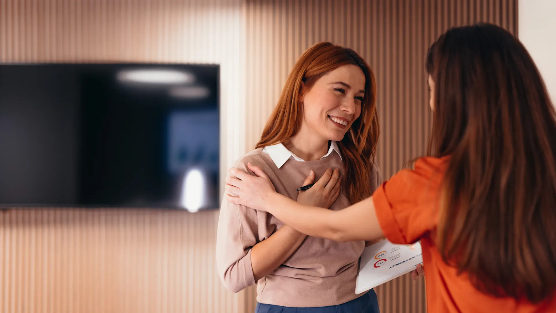 Mentale Gesundheit am Arbeitsplatz (Foto: iStock / Igor Suka) Zwei Frauen begrüßen sich herzlich in einem modern eingerichteten Raum, wobei eine rothaarige Frau in beigem Oberteil lächelnd ihre Gesprächspartnerin in orangefarbener Kleidung die Hand auf die Schulter legt.