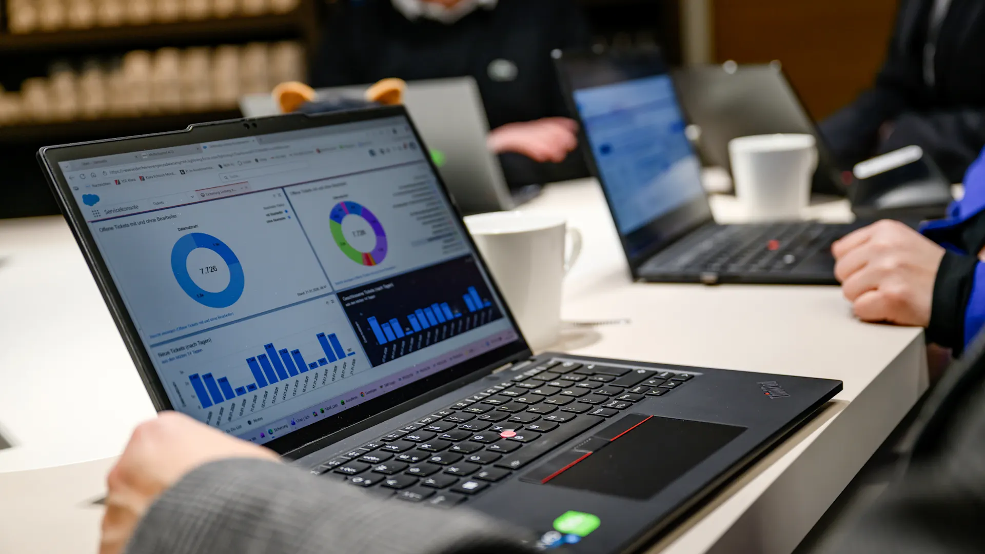 Digitale Ideen, nachhaltige Lösungen. Ein Laptop mit einem Dashboard zur Datenanalyse steht auf einem Besprechungstisch. Im Hintergrund arbeiten mehrere Personen mit weiteren Laptops in einer Besprechung, auf dem Tisch stehen Kaffeetassen.