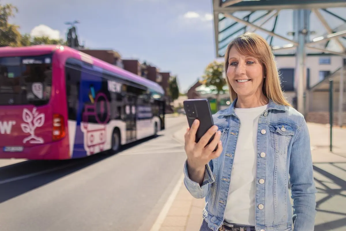 Tickets und Tarife Frau steht an einer Bushaltestelle und schaut in ihr Smartphone. Ein NEW Bus ist im Hintergrund zu sehen.