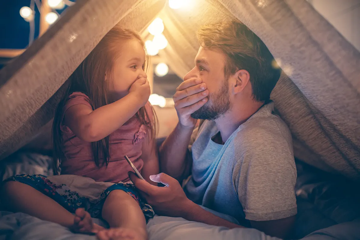 Vater und Tochter freuen sich über günstigen Strom Vater und Tochter sitzen im Wohnzimmer unter einer Deckenhöhle und schauen gemeinsam aufs Smartphone und freuen sich über günstigen Strom.