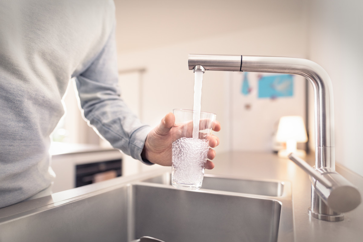Eine Person in hellem Pullover füllt ein Glas mit Wasser aus einem modernen Wasserhahn an einer Küchenspüle. Die Küche hat eine helle, moderne Ausstattung mit Edelstahlelementen und im Hintergrund sind Küchengeräte zu erkennen.