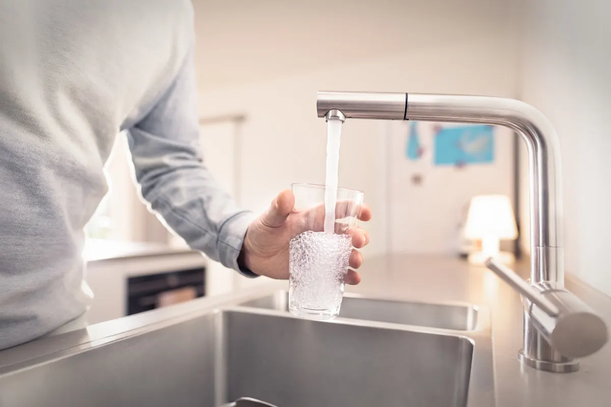 Ressourcen bewahren, Wasser schützen. Eine Person in hellem Pullover füllt ein Glas mit Wasser aus einem modernen Wasserhahn an einer Küchenspüle. Die Küche hat eine helle, moderne Ausstattung mit Edelstahlelementen und im Hintergrund sind Küchengeräte zu erkennen.
