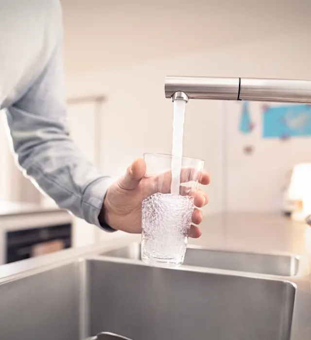 Ressourcen bewahren, Wasser schützen. Eine Person in hellem Pullover füllt ein Glas mit Wasser aus einem modernen Wasserhahn an einer Küchenspüle. Die Küche hat eine helle, moderne Ausstattung mit Edelstahlelementen und im Hintergrund sind Küchengeräte zu erkennen.