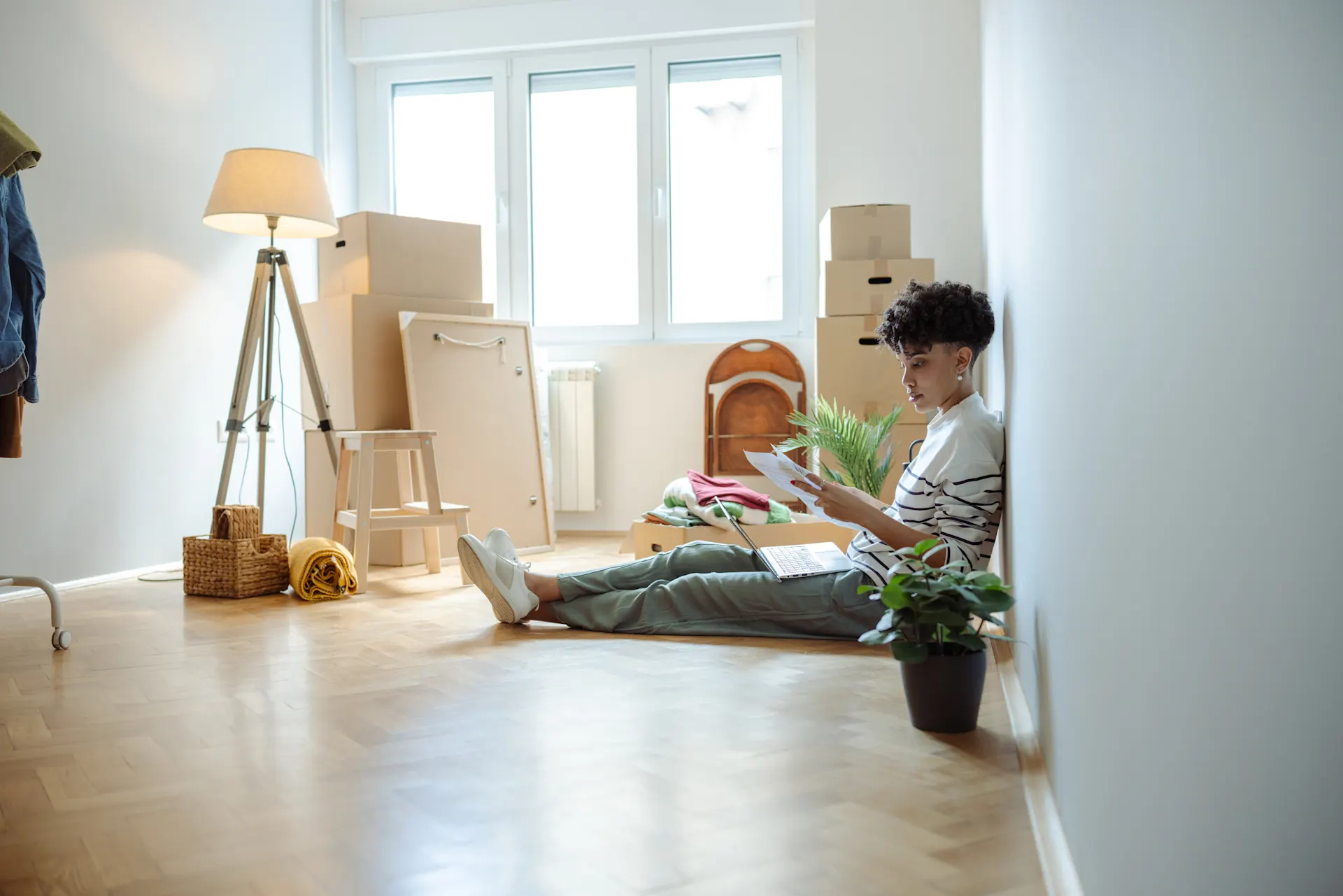 Eine Person sitzt auf einem Holzboden in einem Zimmer mit Umzugskartons, liest Dokumente und hat einen Laptop auf dem Schoß.  