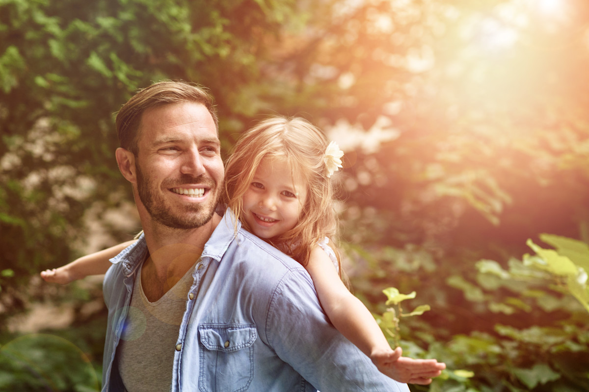 Vater mit Kind auf dem Rücken und Bäumen und Sonne im Hintergrund.