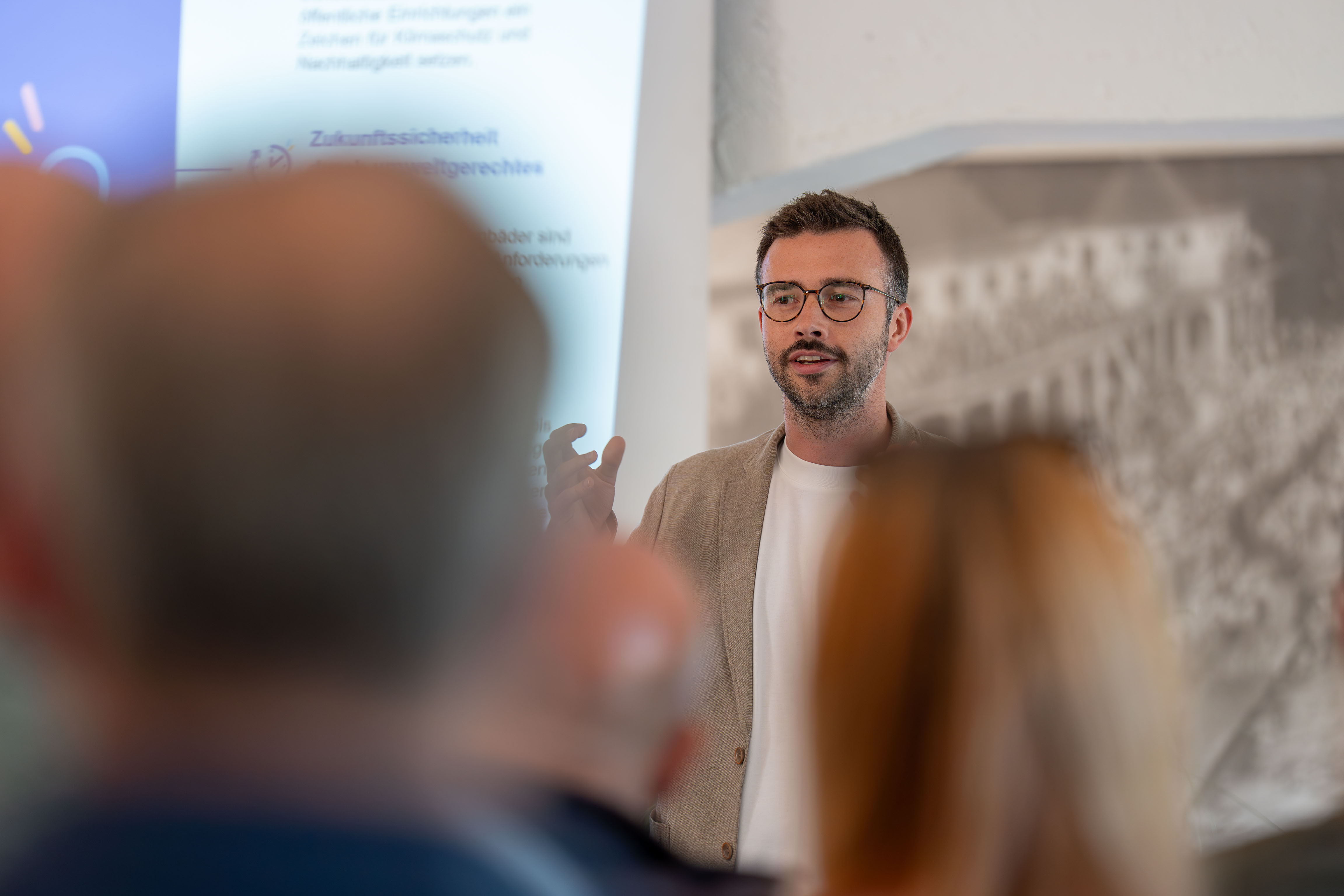  Eine Person mit Brille und beigem Sakko hält einen Vortrag vor Publikum. Die Zuhörenden sind im Vordergrund unscharf zu sehen, während im Hintergrund eine Präsentationsfolie projiziert wird. 

 